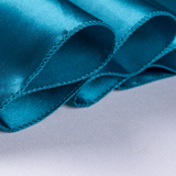 A close-up of teal blue satin fabric with a stitched border, draped over a light gray textured surface.