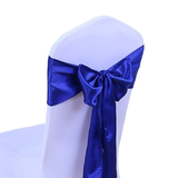A royal blue satin chair sash tied in a large bow and long tail, shown wrapped around a white-covered chair against a plain white background.