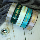 Five rolls of satin ribbon in silver, dark green, mint, teal, and champagne pink, each printed with the words 'Exclusive love you' in gold foil, arranged on a white wooden surface with a gray textured fabric and a small disco ball prop.