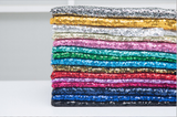A stack of sequined fabric rolls in multiple colors, including silver, gold, white, light blue, pink, green, teal, orange, red, purple, and dark blue, arranged vertically on a white surface.