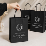 A hand holds a black paper gift bag with black handles in a studio setting. The bag has a white printed monogram logo reading CJ & J, a wreath, and the text THANK YOU Caroline & Johnson with the date 06.