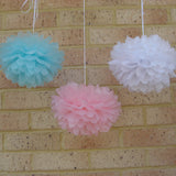 Three tissue paper pom-poms in light blue, pink, and white, hanging against a tan brick wall.
