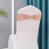 A white chair cover decorated with a metallic rose gold sash tied in a bow at the back, featuring a jeweled ring accent, set against a background of gray walls and teal curtains.