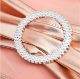A circular silver-toned ring embellished with small clear rhinestones, displayed on a rose gold satin ribbon against a matching peach background.