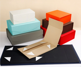 A set of colorful rectangular boxes and a tan folding box displayed on a white surface against a beige background. The boxes come in white, light blue, dark gray, orange, brown, red, and black, with one tan box shown open in front.
