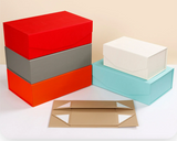 A set of flat cardboard packaging boxes shown on a clean white surface against a light beige background. The set includes three stacked rectangular boxes in red, gray, and orange, a single white box, a single light blue box, and one flat tan box with printed white corner triangles.