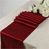 A deep red satin table runner draped diagonally across a white tablecloth, styled with white roses at the top right.