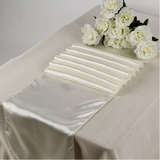 An ivory satin table runner draped diagonally across a table covered with a matching ivory tablecloth, styled with white artificial roses in the corner.