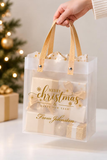 A clear plastic gift bag with gold handles and gold holiday lettering reading 'Merry Christmas & Happy New Year! From Johnston,' held by a hand in front of a softly blurred Christmas tree and wrapped presents.