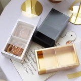 Three rectangular gift boxes arranged on a white tablecloth with printed paper and gold decorative objects. The boxes include a white marble-patterned box with a clear window, a dark gray closed box, and an open gold-toned box showing a beige interior.