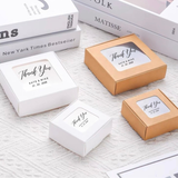 Four small square thank-you gift boxes arranged on a light textured fabric surface, including two white boxes and two tan/golden boxes, each with a clear window and printed personalized text. Books are partially visible in the background.