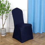A single high-back chair covered in a dark navy blue stretch spandex chair slipcover, shown in a styled room setting with a light wood floor, a potted plant, and a light blue curtain.